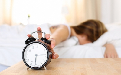 Sleepy young woman trying kill alarm clock while bury face in pillow. Early wake up, not getting enough sleep, getting work concept. Female stretching hand to ringing alarm willing turn it off