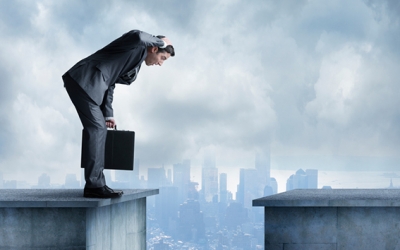 A businessman, holding a briefcase, stands on the ledge of a building. There is another building opposite of him with a gap in between the two. He is leaning over with his mouth open wide and his hand on his head. A big city skyline is in the distance obscured by clouds.